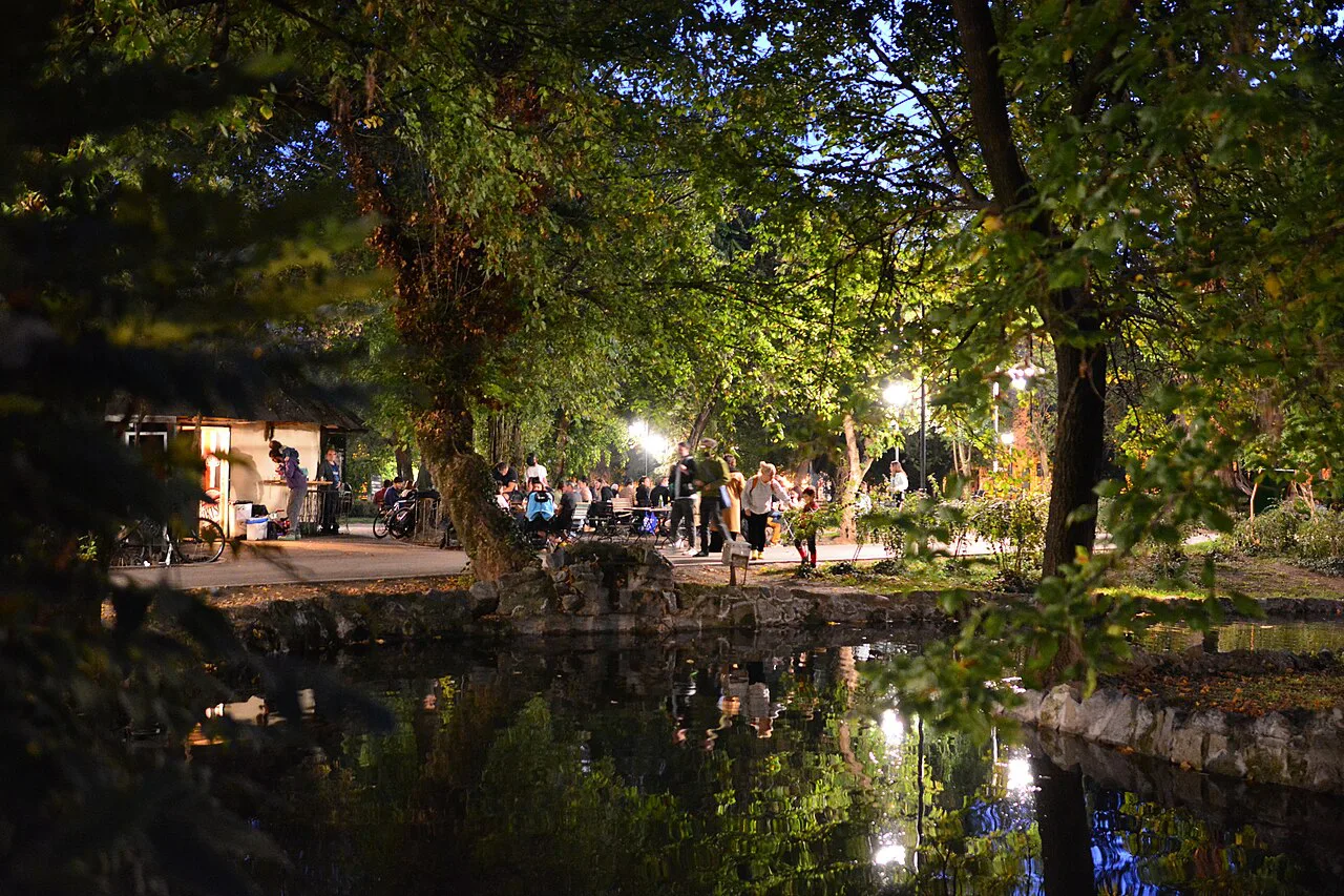An outdoor bar beside a pond in Cișmigiu Gardens, Bucharest, at dusk.